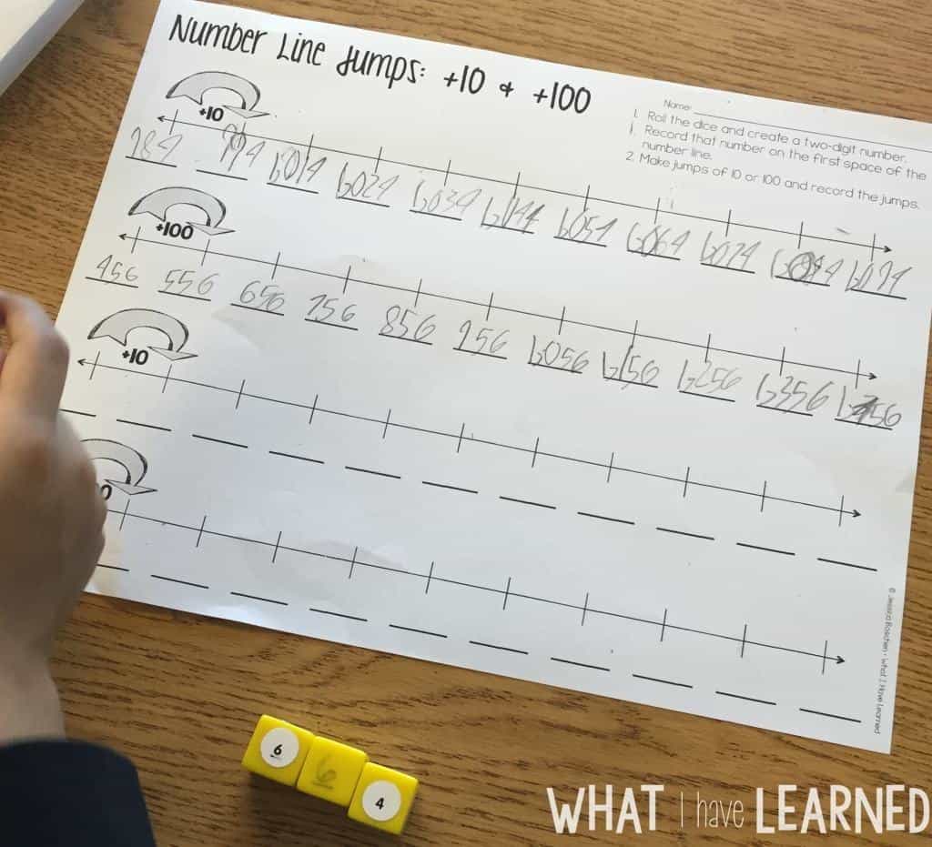 How to Teach a Second Grade Number Line for Addition and Subtraction 8 A child works on a math worksheet titled “Number line jumps: +10 & +100,” drawing number lines and recording jumps by tens and hundreds. Yellow number dice and a pencil are spread out on the table.