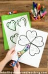 A hand cuts out a shamrock shape from a worksheet listing reasons to feel lucky—a charming St. Patrick's Day craftivity—next to a green folder, heart cutouts, and colorful markers on a wooden table.