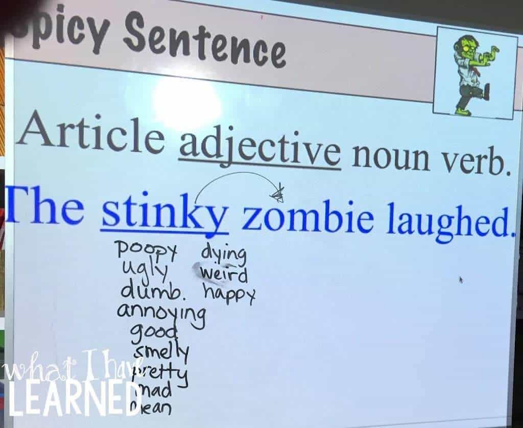 A classroom whiteboard displays an Article adjective noun verb sentence template. Example: The stinky zombie laughed. Below it is a list of adjectives&mdash;poopy, dying, weird&mdash;under this clever paragraph.