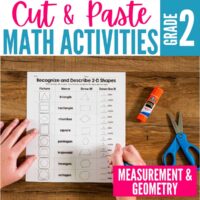 A student’s hands work on a Cut and Paste Math Activities for Second Grade - Measurement and Geometry worksheet, with a glue stick and blue scissors nearby on a wooden table. Text reads: Cut & Paste Math Activities Grade 2.