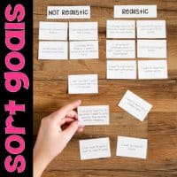 Someone sorts goal cards under Not Realistic and Realistic columns on a wooden table. "Sort Goals" appears vertically. Shown: Are you SMART? Goal Setting Lessons.