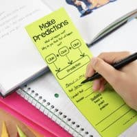 A hand writes on a green “Make predictions” worksheet inspired by the Reading Comprehension Bookmarks, encouraging students to use clues to predict outcomes. The worksheet sits atop open notebooks with colored pencils scattered nearby.