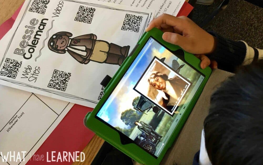 Using QR Codes in the Classroom1 A student uses a tablet to watch a Bessie Coleman video, with the "Using QR Codes in the Classroom1" worksheet—featuring Bessie Coleman illustrations and QR codes—on the desk. “WHAT have I LEARNED” prompts reflection using the QR codes.