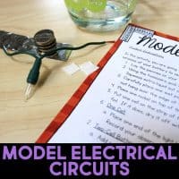 A simple electrical circuit project with stacked coins, foil, a small bulb with attached wires, and a glass of water next to a printed instruction sheet titled “Model Electrical Circuits” on a wooden surface.