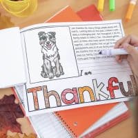 A child colors the Thankful Craftivity Flap Book, which features a dog drawing and writing lines. The flap book is on orange notebooks, with autumn leaves nearby.