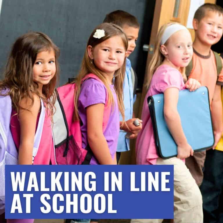 Five young children with backpacks stand walking in a line inside a school, smiling at the camera. Text overlay reads “WALKING IN LINE AT SCHOOL.”.