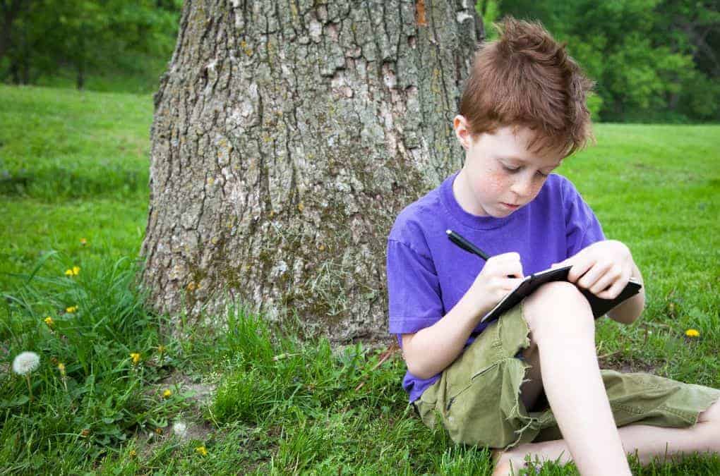 A young boy with reddish hair, wearing a purple T-shirt and green shorts, sits on grass in a park, leaning against a tree and writing or drawing different types of letters in a notebook on his lap.