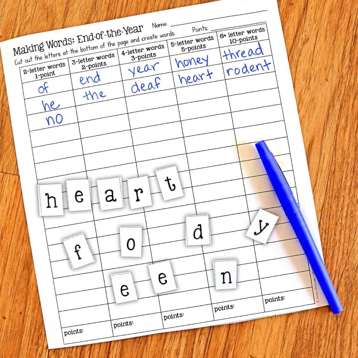 A worksheet titled Making Words: End-of-the-Year lies on a wooden surface with a blue pen and letter tiles arranged to spell heart, food, and eye. The worksheet lists words and their point values.