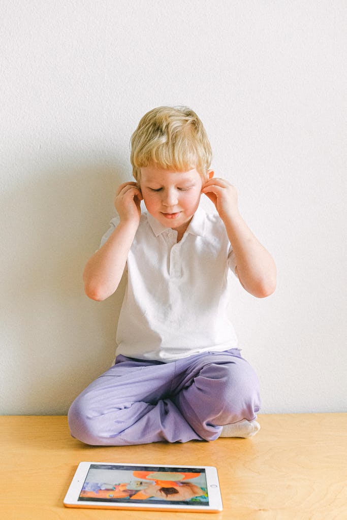 A cute blond child sits on a wooden floor indoors playing with a tablet, creating an adorable tech-savvy moment.