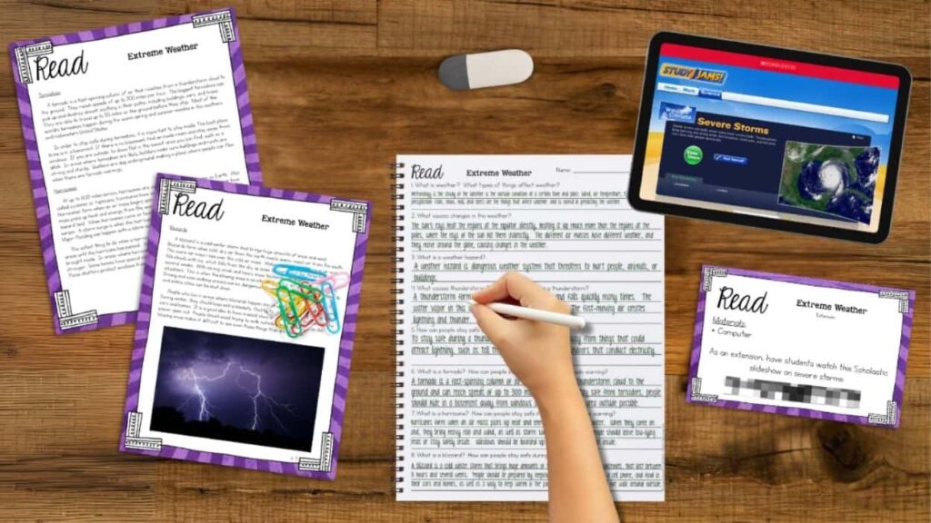 A hand writes in a notebook on a wooden desk surrounded by PREVIEW 2715136 READ about Extreme Weather - Weather & Climate - 3rd Grade _1 materials, science station reports, paper clips, and a tablet showing weather data with a Severe Storms headline.